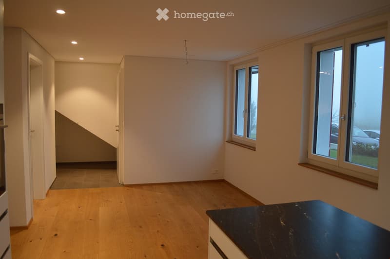Empty room with light wooden flooring, white walls, and large windows with wooden sills. The room features recessed lighting and a small black countertop in the foreground.