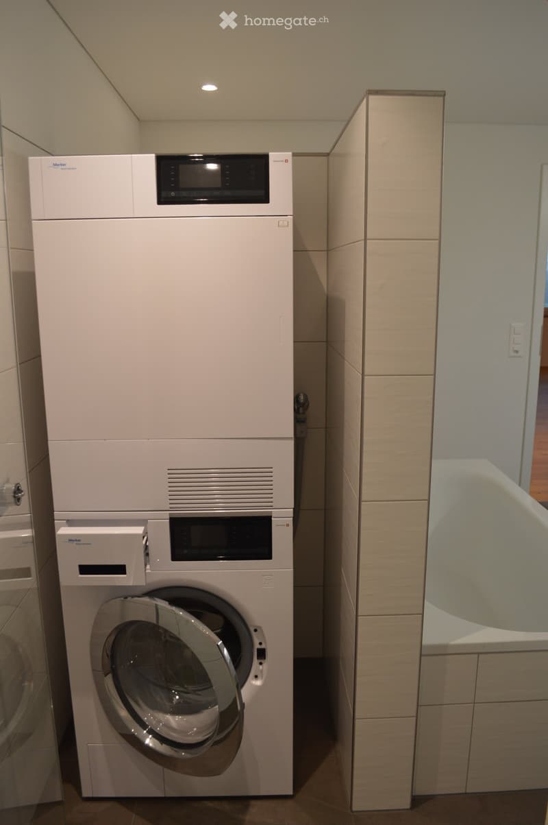 Modern bathroom with a white washing machine and dishwasher stacked vertically, sleek design, light-colored tiles, and a small window.