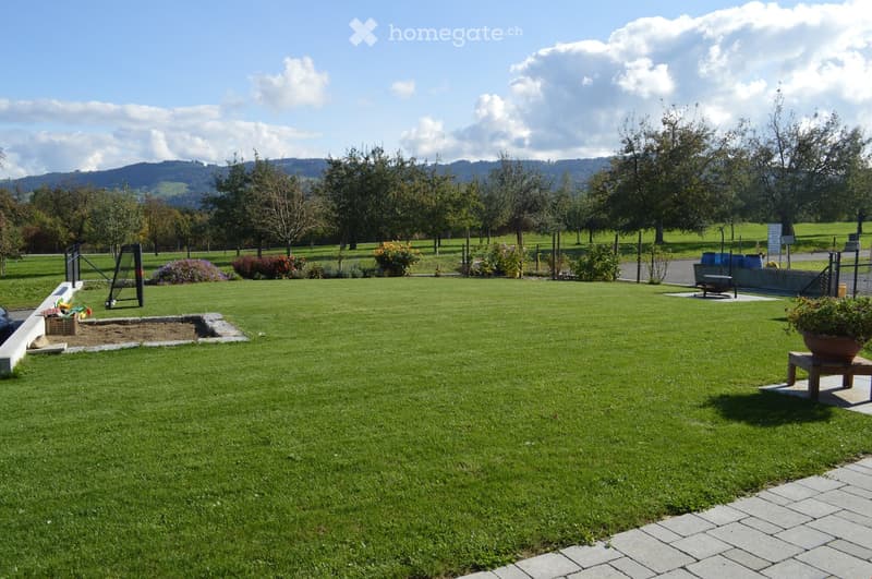 Well-maintained lawn with a small sandbox, a few potted plants, and a bench, surrounded by trees and a fence with a scenic mountain backdrop.