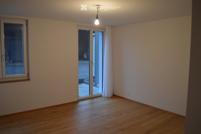 Empty room with wooden flooring, white walls, large window, and sliding glass door leading outside.