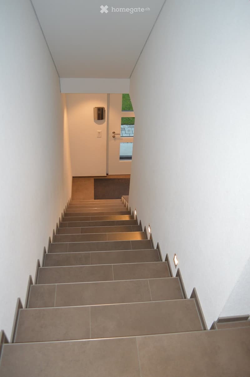 Modern staircase with beige tiled steps and walls, illuminated by wall-mounted lights, leading to a door with a small window showing greenery outside.
