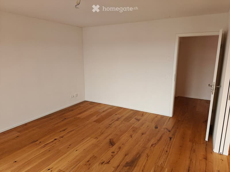 Empty room with light-colored walls and wooden flooring, featuring an open doorway leading to another room.