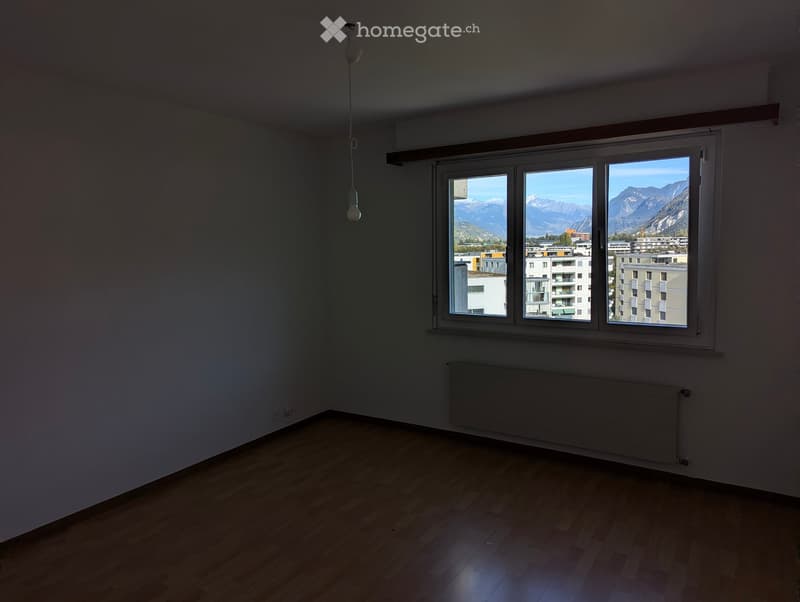 Empty room with wooden flooring, large window showing a view of mountains and buildings, radiator, and a hanging light fixture.