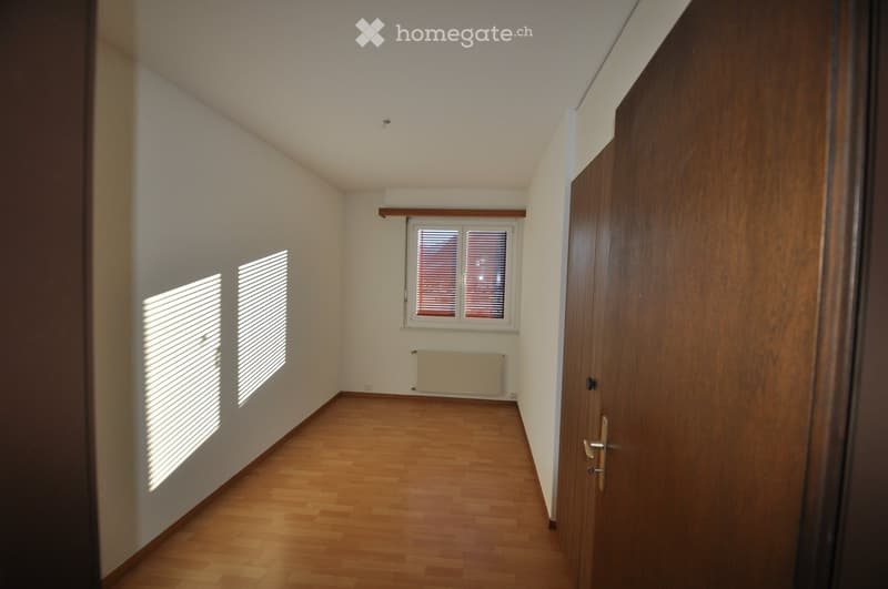 Empty room with wooden flooring, white walls, and two windows with blinds. A radiator is visible below one window, and a wooden door is on the right side.