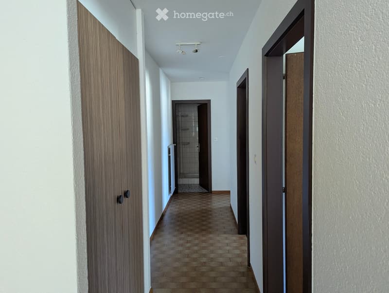 A narrow hallway with tiled flooring, white walls, and several wooden doors leading to different rooms, including a visible bathroom.