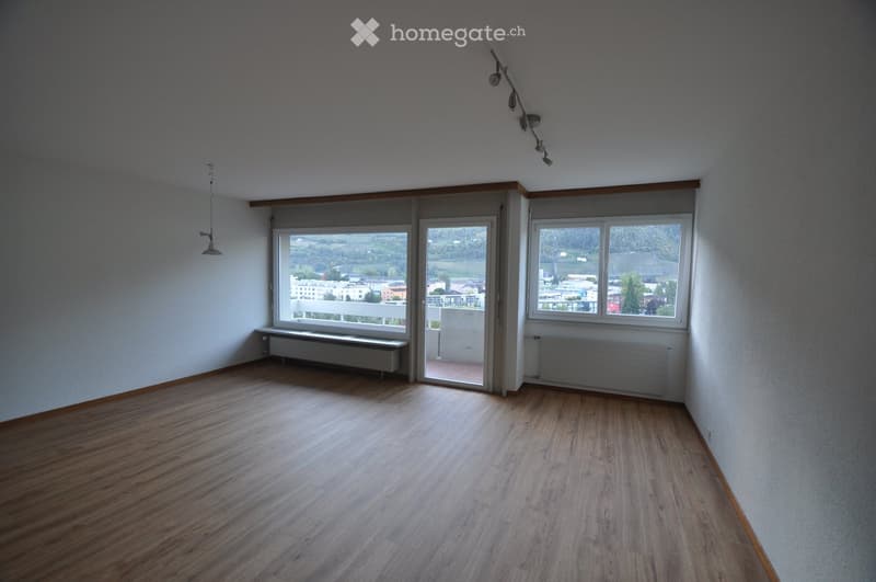 Empty room with wooden flooring, white walls, large windows, and a sliding glass door leading to a balcony.
