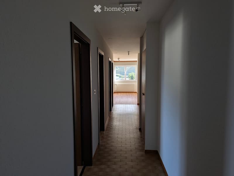 A long, narrow hallway with tiled flooring, wooden doors on both sides, and a view of a room with wooden flooring and large windows at the end.