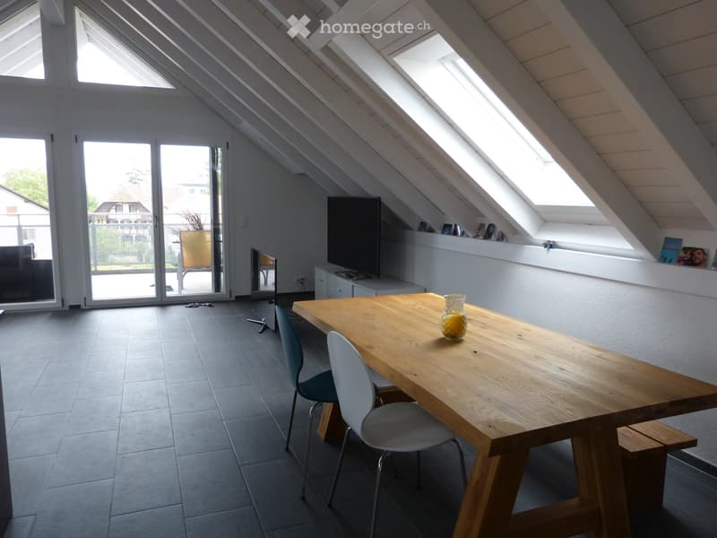 Bright attic room with wooden table, chairs, TV, and large windows, featuring a modern design and ample natural light.