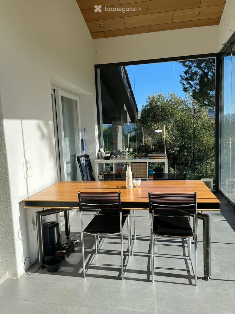 Balcony with a wooden table, two chairs, a bottle, and a cup, overlooking a scenic view with trees and mountains.