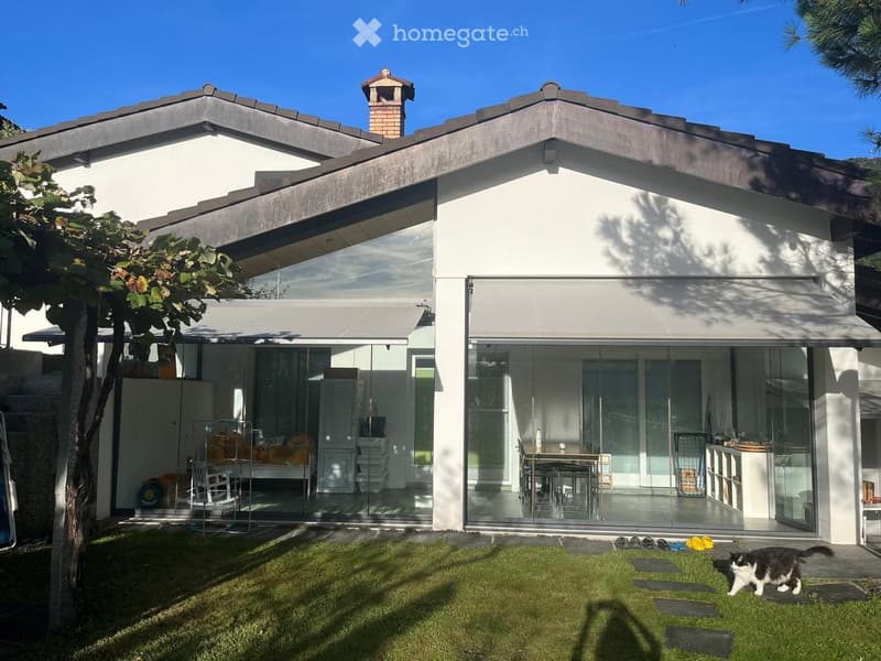 Modern two-story house with white exterior, large glass sliding doors, and a well-maintained lawn. The house features a raised ground floor and a small chimney on the roof.