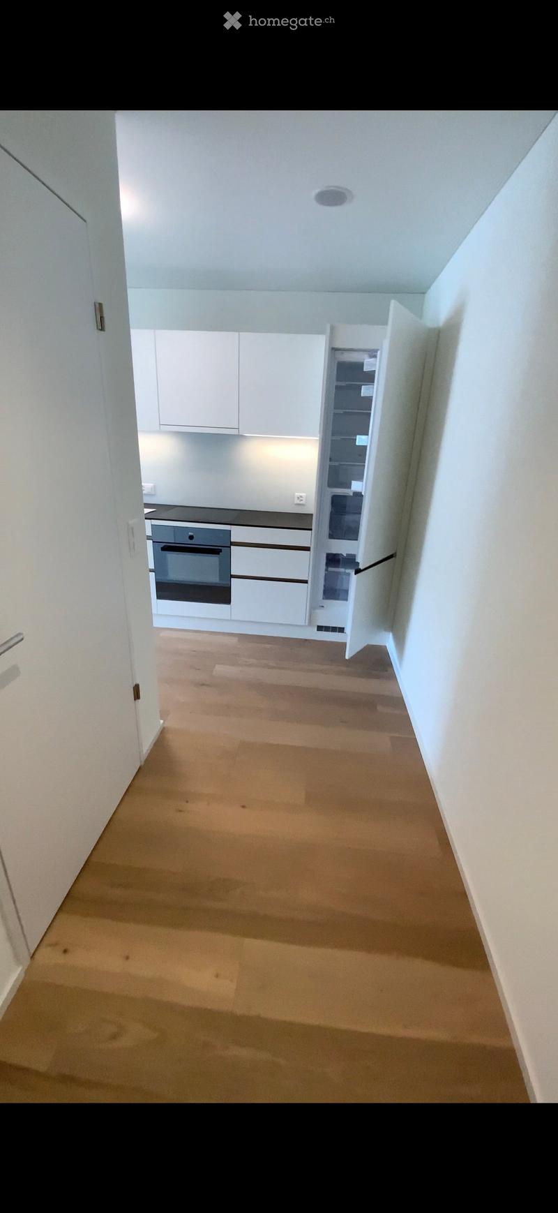 Empty hallway with wooden flooring leading to a modern kitchen area featuring white cabinets, a built-in oven, and a refrigerator. The kitchen is clean and unfurnished.