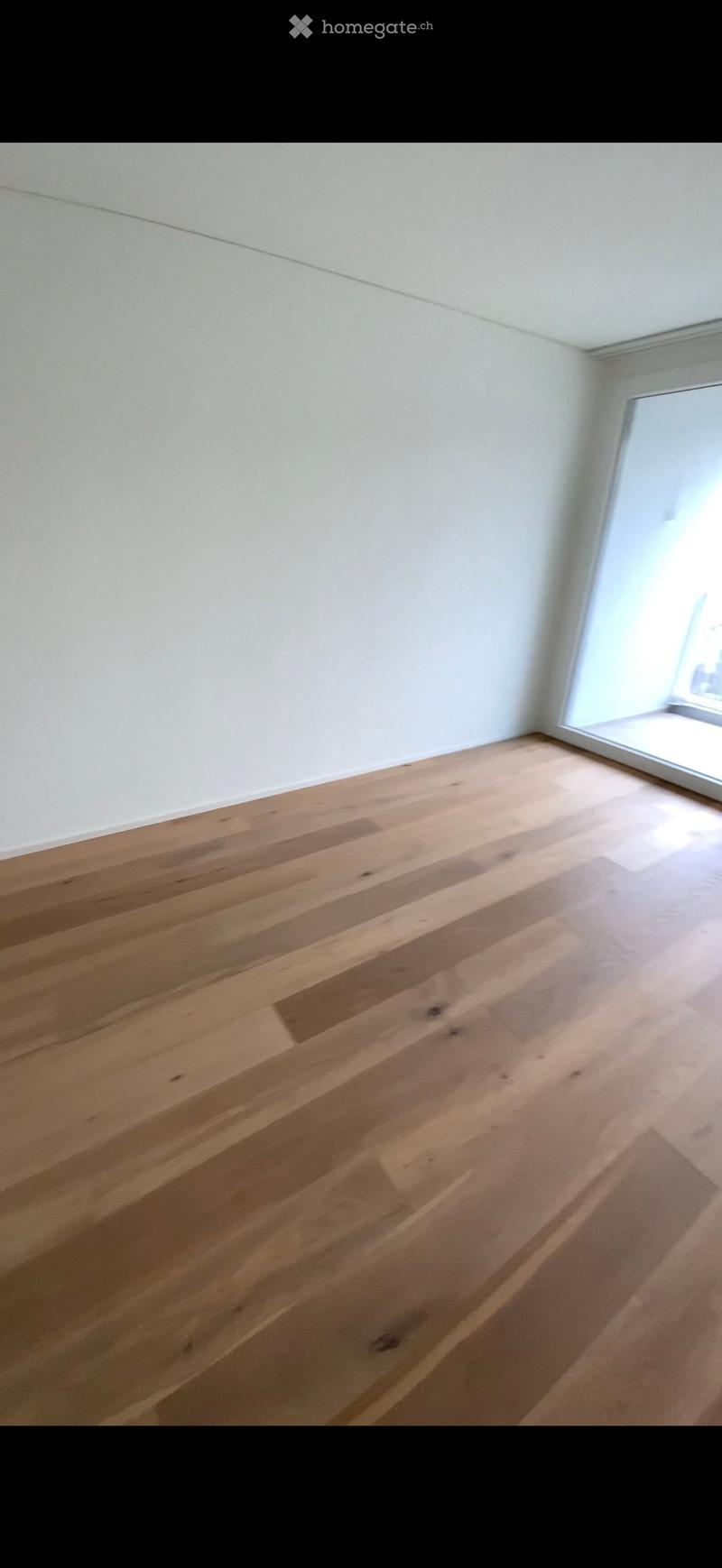 Empty room with light wooden flooring and white walls, featuring a large window on one side.