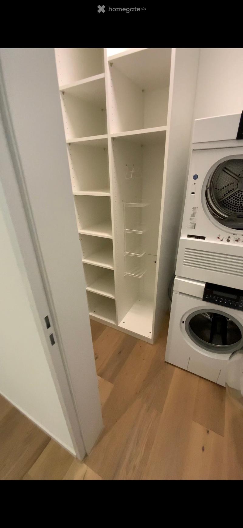 Empty room with wooden flooring, white built-in shelving unit, and a white washing machine visible in the corner.