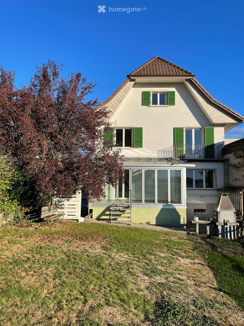 Two-story house with white walls, green shutters, and a tiled roof. Features a balcony on the upper level and a patio area with a barbecue and seating.