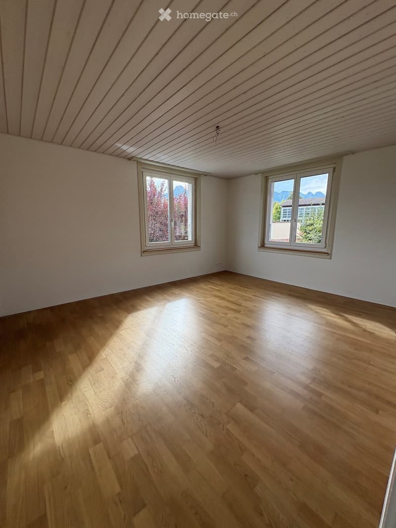 Empty room with wooden flooring, white walls, and two windows offering a view of the outdoors.