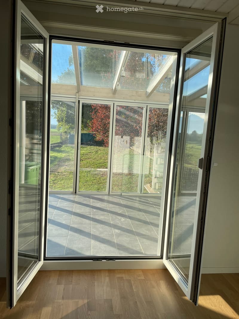 Bright, empty room with wooden flooring and large glass doors leading to a patio area with tiled flooring and outdoor view.