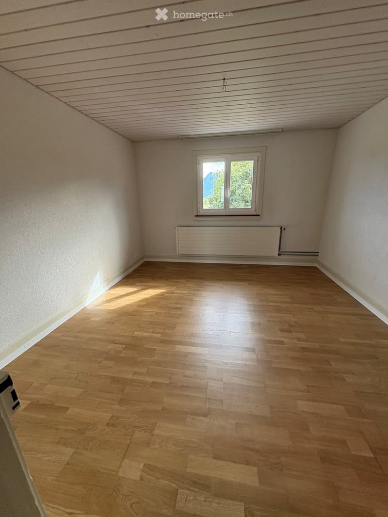 Empty room with wooden flooring, white walls, and a single window letting in natural light. A radiator is mounted on the wall below the window.