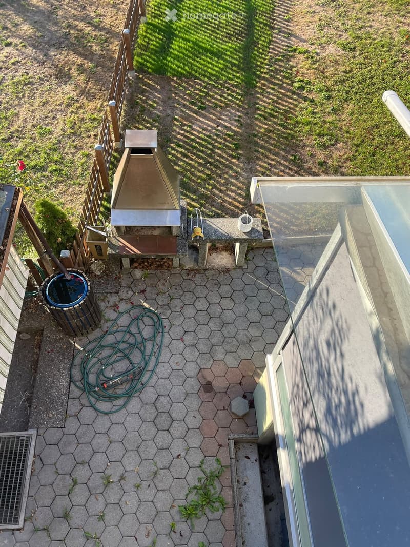 A small patio area with hexagonal tiles, a wooden fence, a small shed, a hose, a trash bin, and a small pool with a glass cover.