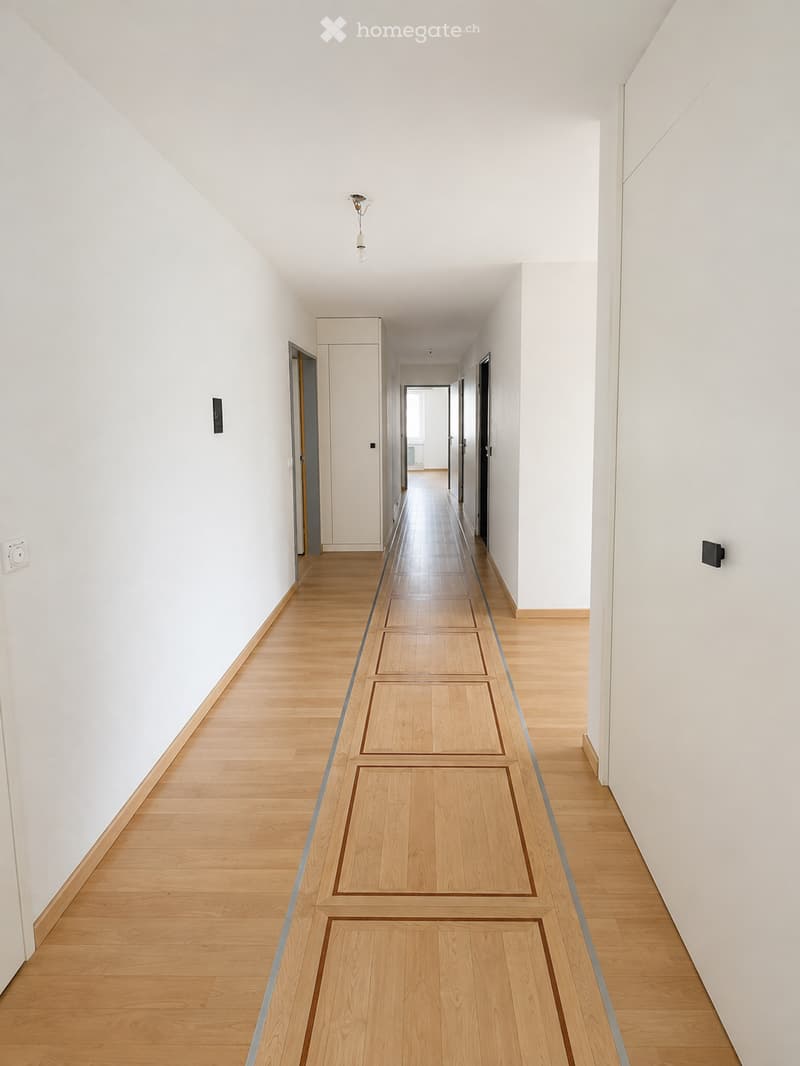A clean, modern hallway with light wooden flooring and white walls, featuring several doors and a single ceiling light fixture.