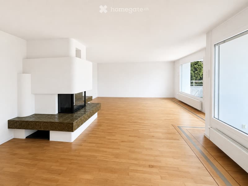 Empty room with light wooden flooring, white walls, and a built-in fireplace with a decorative tiled surround. Large windows provide natural light.