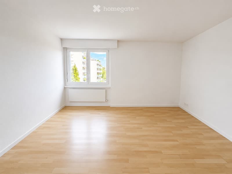 Empty room with light wooden flooring, white walls, and a large window with a view of trees and buildings outside.