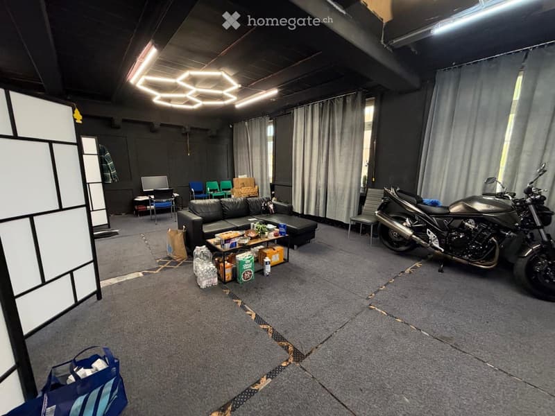 A spacious, modern room with a grey carpeted floor, a black sofa, a small table with snacks, a screen, and a motorcycle parked in the corner.