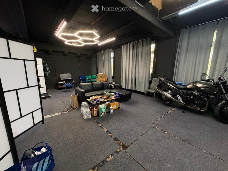 A spacious room with a grey carpeted floor, a black leather sofa, a small table with snacks, a motorcycle, and several chairs arranged around the space.