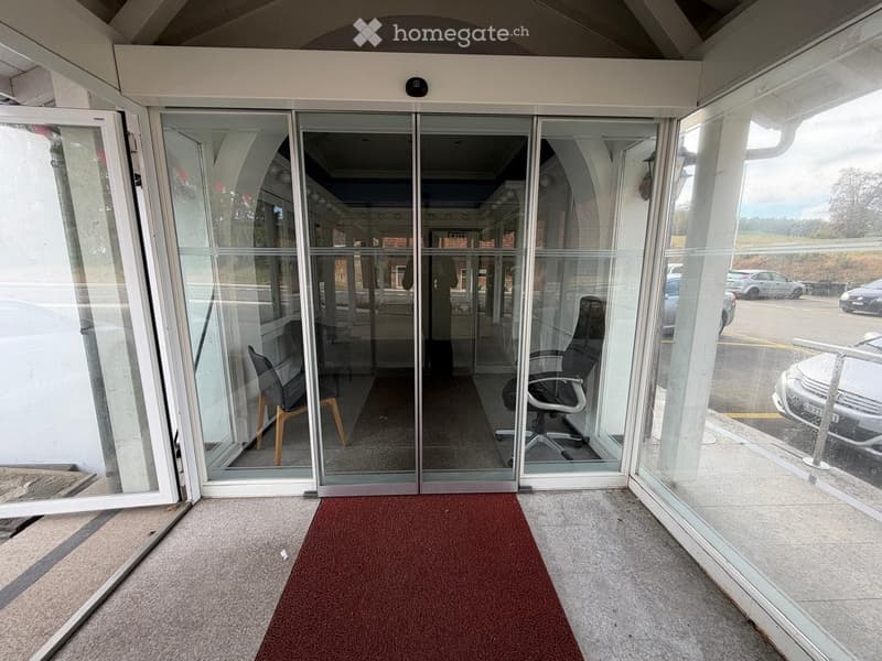 Entrance area with automatic glass doors, red carpet, and visible office chairs inside the building.