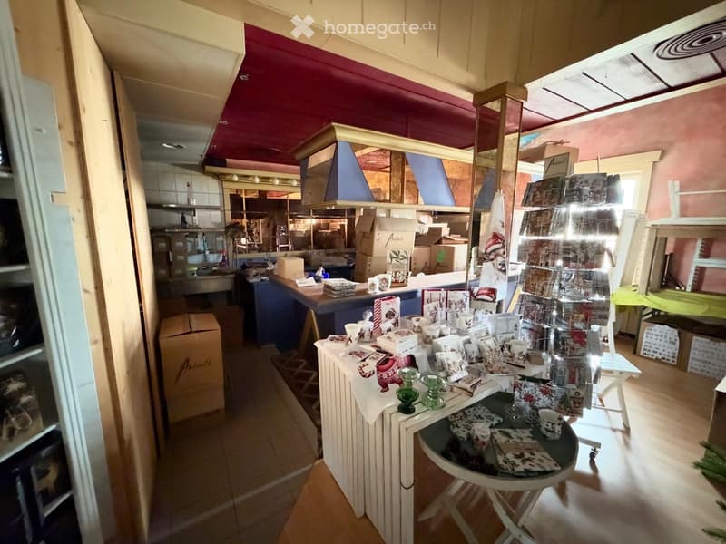 A small shop or store with a counter, display racks, and a kitchen area in the background. The counter has cups, a small table, and decorative items.
