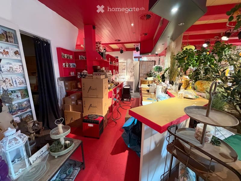 A brightly lit shop with red walls and ceiling, featuring shelves filled with various items, including cakes, cups, and decorative items. There are tables and stands displaying cakes and other products.