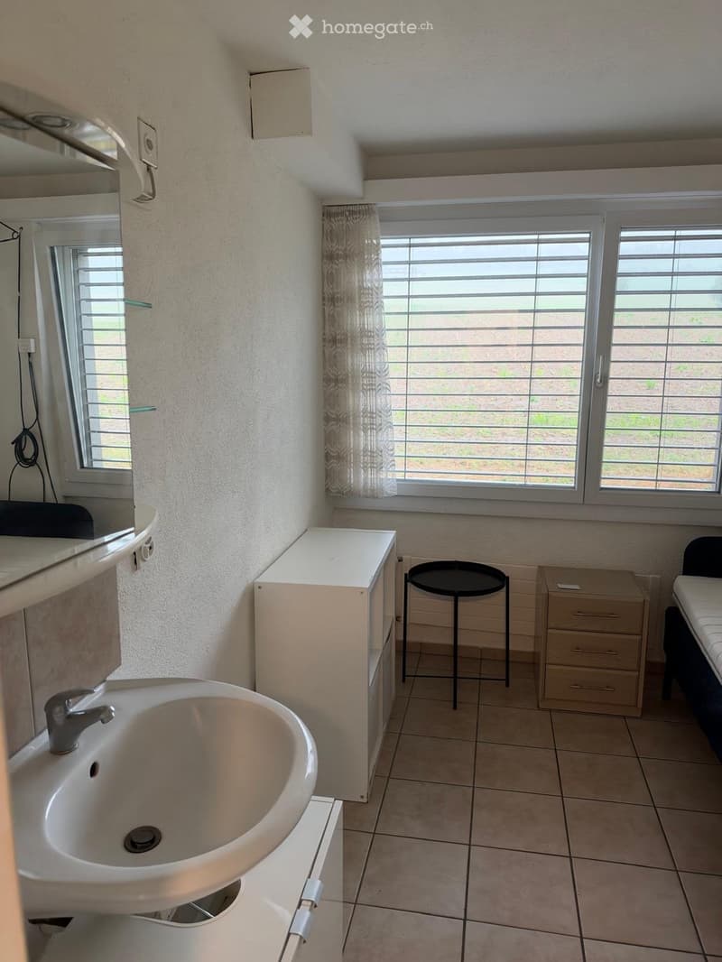 A small, furnished room with a sink, a stool, a cabinet, a drawer unit, and a curtain on the window.