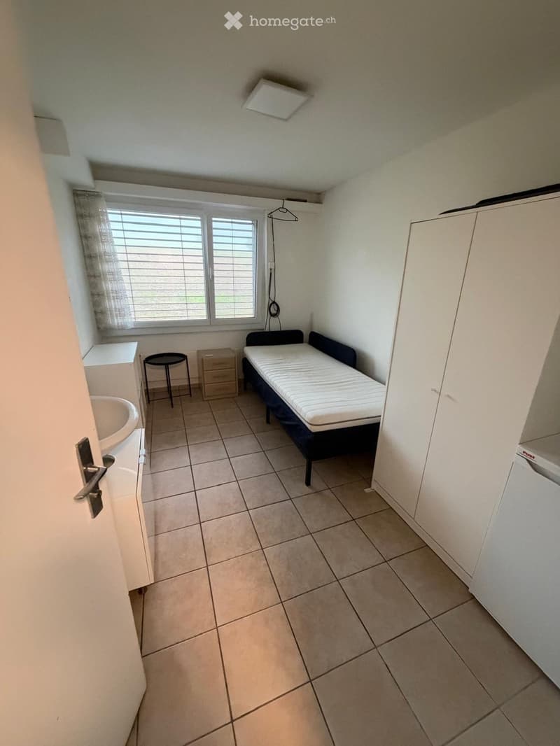 A small, furnished room with a bed, a stool, a cabinet, a sink, and a drawer. The room has tiled flooring and a window with blinds.