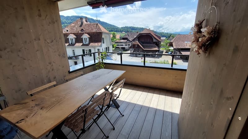 Balcony with wooden table and chairs, overlooking a village with traditional houses and mountains.