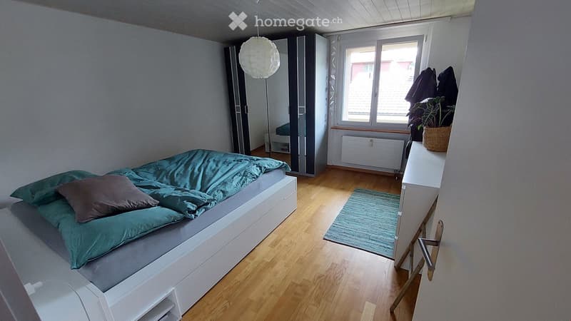 Modern bedroom with a white bed, teal bedding, a nightstand, a wardrobe, a hanging lamp, a small plant on a shelf, and a rug on the wooden floor.