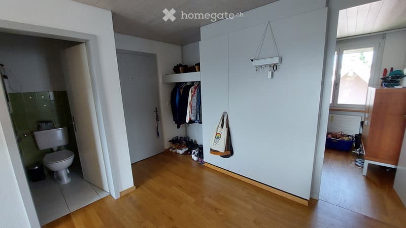 The image shows a hallway with wooden flooring, a white wardrobe with a coat rack, a shelf with shoes, a handbag hanging on the wall, a radiator, and a cabinet. A bathroom is visible to the left with a toilet and a tiled wall.
