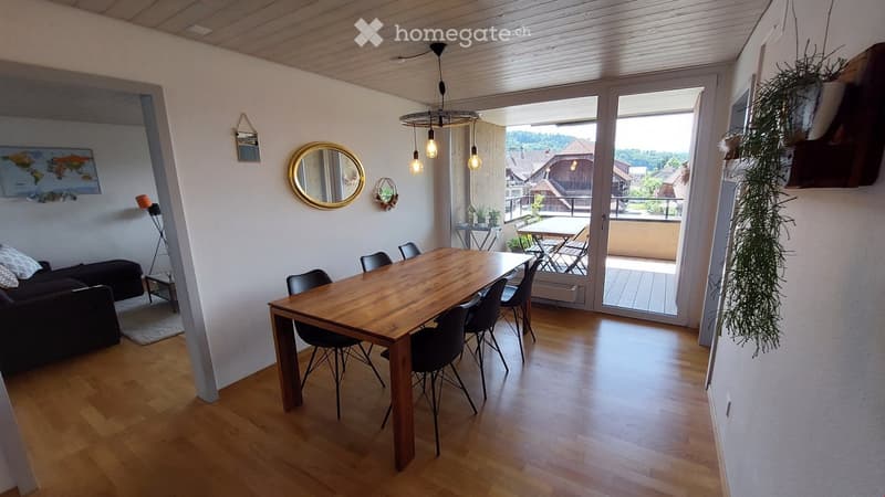 Bright living and dining area with wooden flooring, a large wooden dining table with chairs, a sofa, a hanging pendant light, and a balcony with outdoor furniture visible through large glass doors.