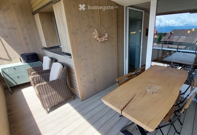 Balcony with wooden floor, wicker chair, wooden table, and view of the surrounding area.
