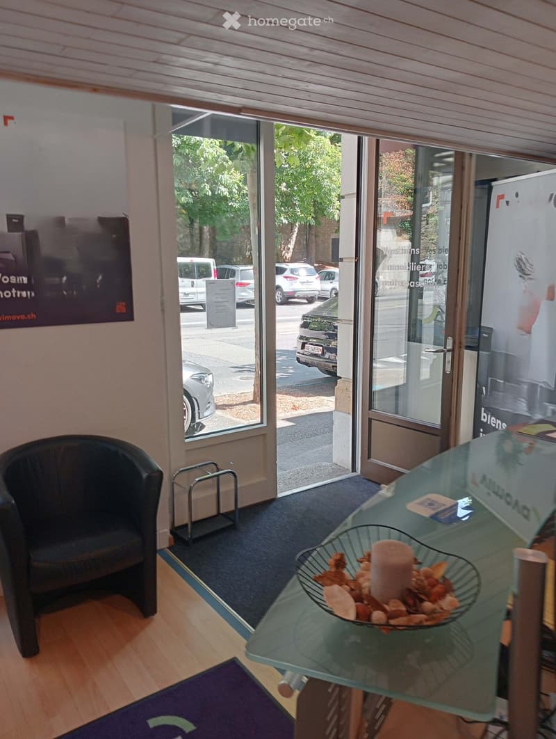 A small, well-lit office or reception area with a wooden floor, a glass table with a decorative bowl, a single chair, and a small metal stand near the entrance.