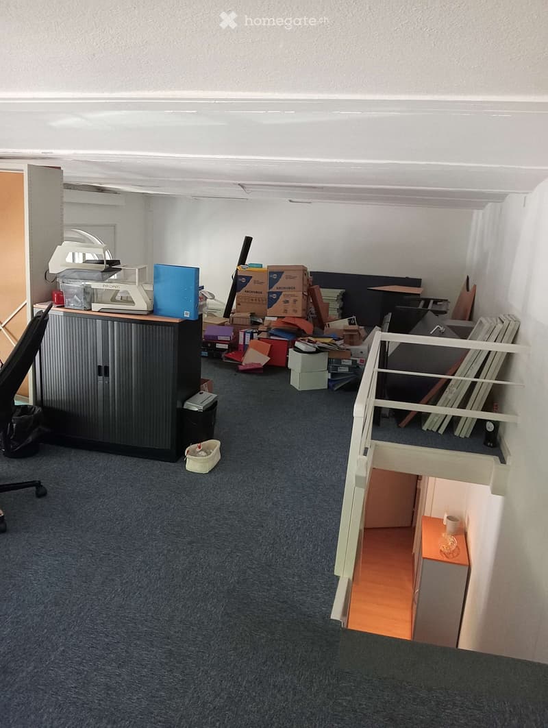 The room has a grey carpeted floor, white walls, and a black cabinet with a printer on top. There is a small white stool with a cup on it, a black chair, and several boxes and items scattered around. A small staircase leads to another area with a small ta