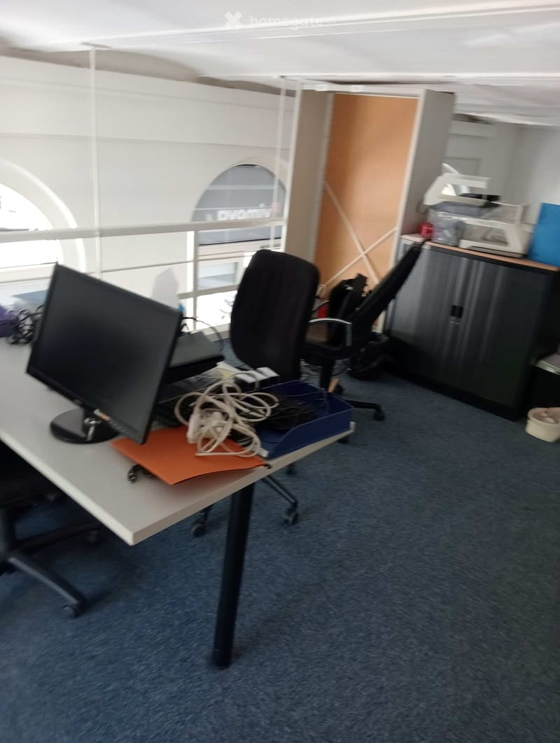 Office space with a desk, chair, screen, printer, and cabinet. Large windows provide natural light.