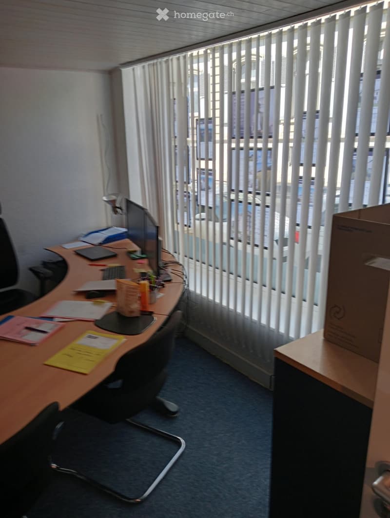 Office space with a wooden desk, office chair, laptop, screen, mouse, keyboard, and papers on the desk. There is a cardboard box on a small table to the right.