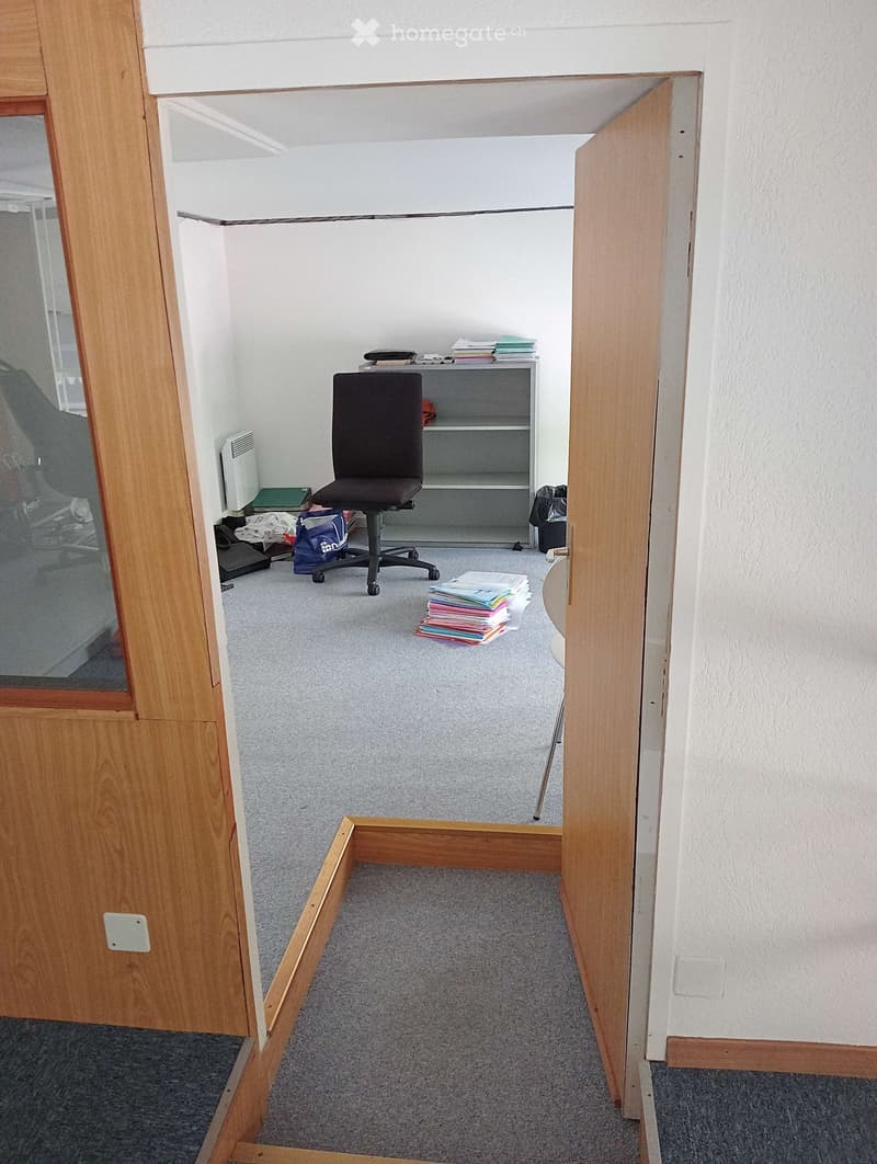 Small office space with a grey carpet, a black office chair, a small shelf, and a round table with papers.