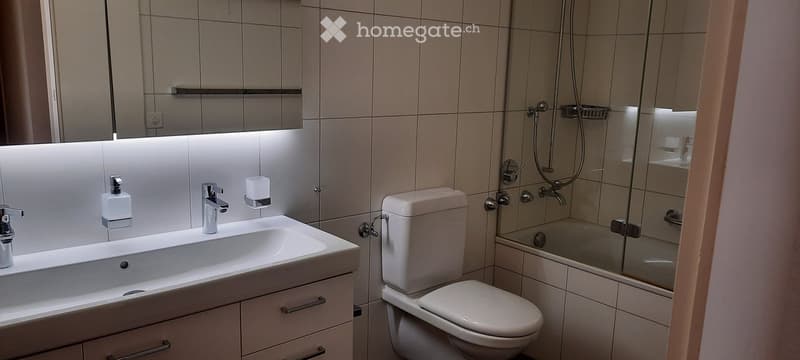 Modern bathroom with a white sink, toilet, and glass-enclosed shower. Features a large mirror with built-in lighting and a sleek faucet.