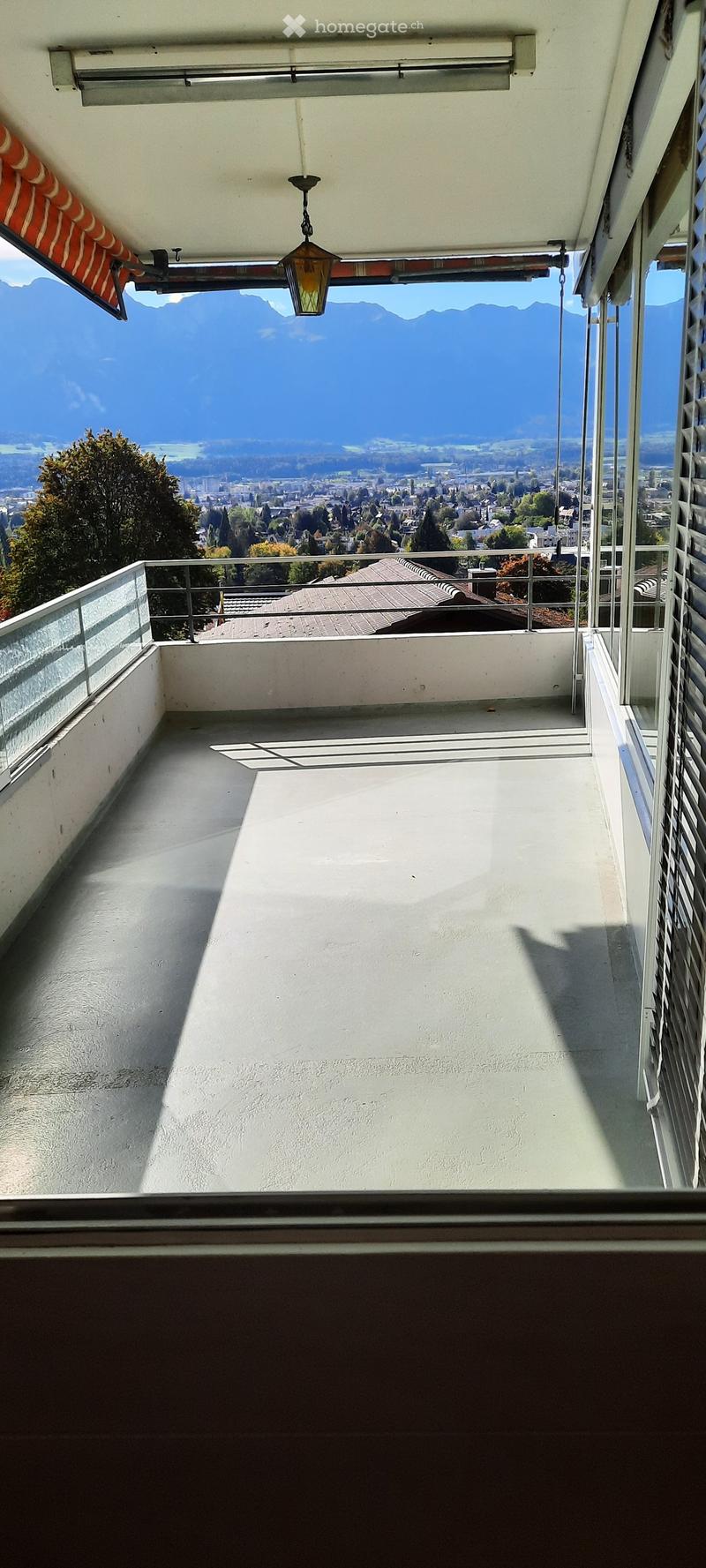 Balcony with a panoramic view of a city and mountains, featuring a glass railing and a hanging light fixture.