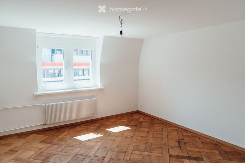 Empty room with wooden herringbone flooring, white walls, large window with white frame, radiator below window, and a single light fixture hanging from the ceiling.