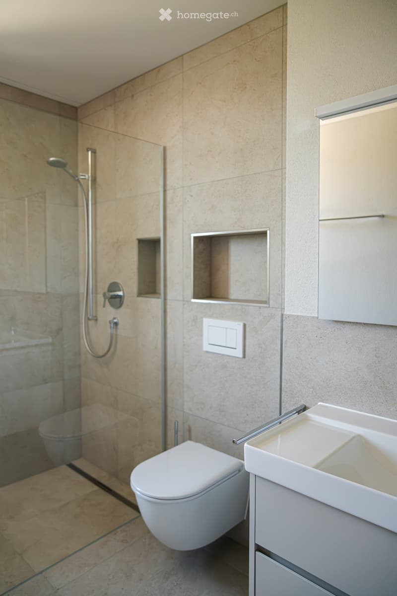 Modern bathroom with beige tiles, a white toilet, a sleek sink with a modern faucet, and a glass-enclosed shower area with a handheld showerhead.