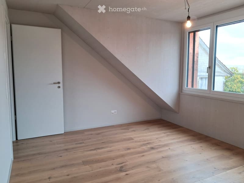 Empty attic room with wooden flooring, white walls, and a large window offering a view of the outside. The room features a slanted ceiling and a single door.
