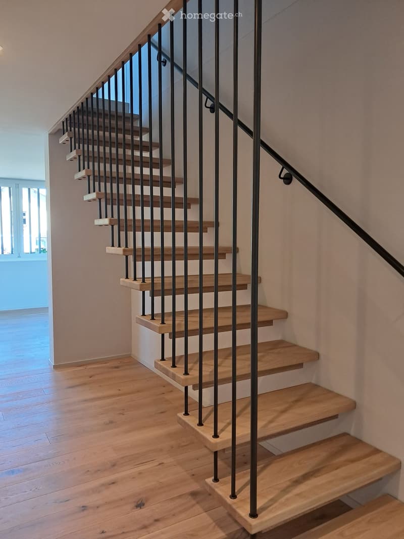 Wooden staircase with black metal balustrade, light wooden steps, and a handrail, set against light-colored walls and wooden flooring.