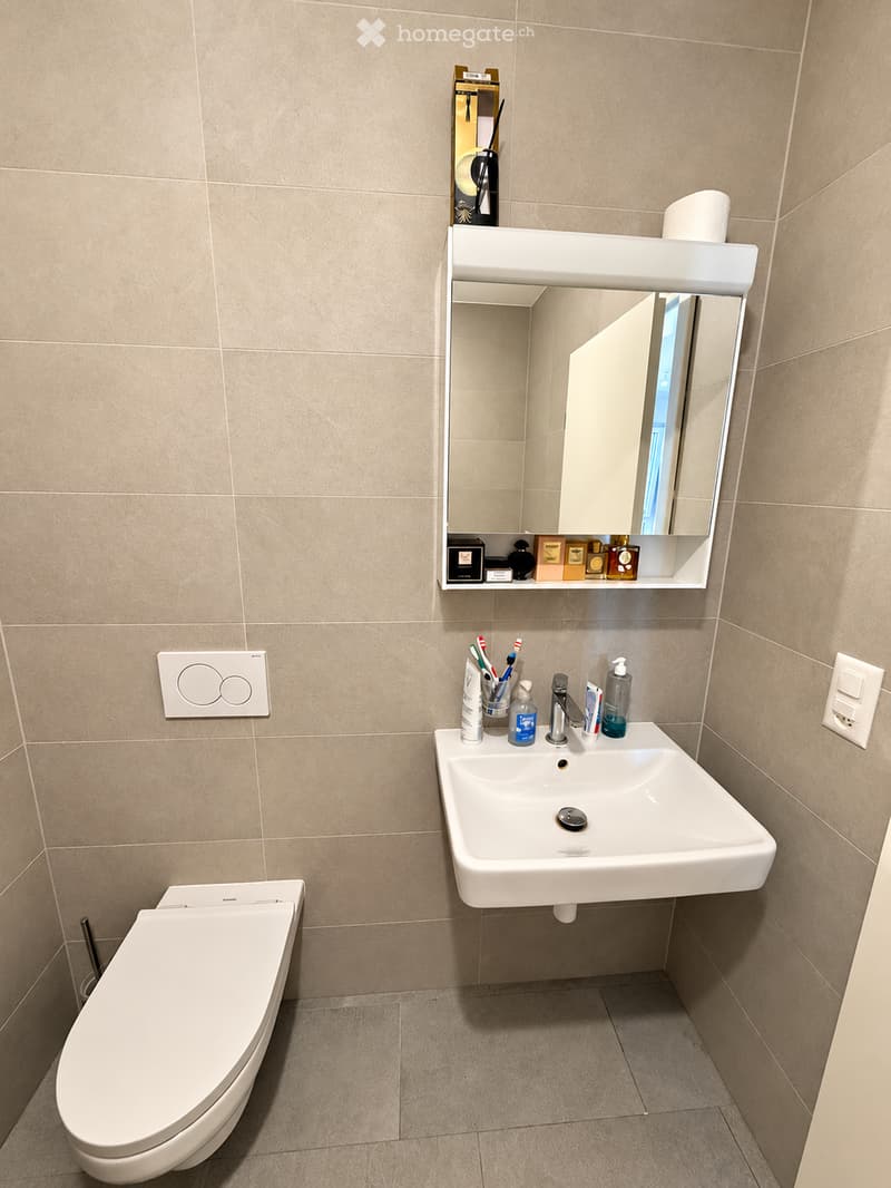 Modern bathroom with beige tiled walls and floor, white toilet, sink with mirror above, and various toiletries on the sink counter.