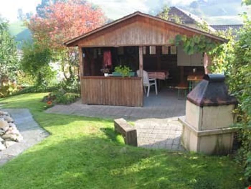 A wooden outdoor structure with a seating area, surrounded by a well-maintained lawn and trees.